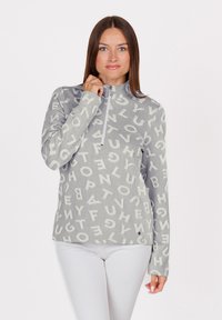 Donna con lunghi capelli castani che indossa un top grigio a maniche lunghe con lettere bianche sparse e pantaloni bianchi, mentre aggiusta il colletto con la zip del top.