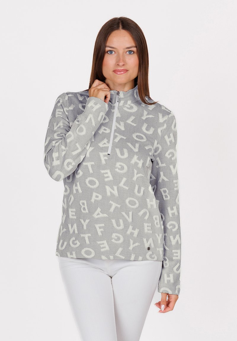 Donna con lunghi capelli castani che indossa un top grigio a maniche lunghe con lettere bianche sparse e pantaloni bianchi, mentre aggiusta il colletto con la zip del top.