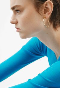 Pendiente de aro de oro combinado con una blusa ajustada de manga larga azul. El fondo es blanco, lo que resalta los colores de la joyería y la prenda.