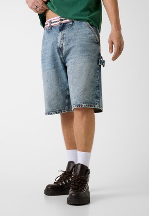 Osoba ubrana w wyblakłe krótkie jeansy, zieloną koszulę, białe skarpety i ciemnobrązowe buty trekkingowe, stojąca na jednolitym tle.