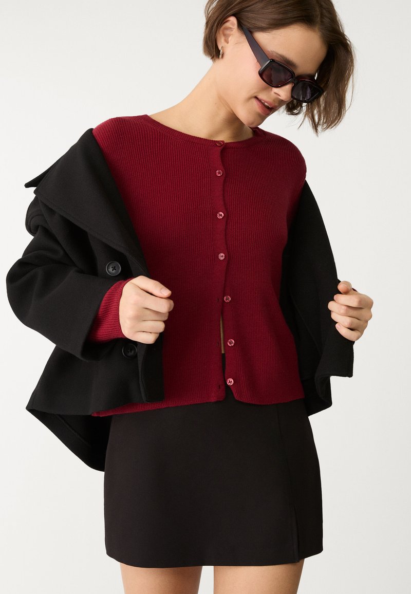 Ribbeliger kastanienbrauner Pullover mit sechs Knöpfen, unter einem schwarzen Mantel getragen, kombiniert mit einem schwarzen Rock. Glatte Textur, lässige Passform und maßgeschneiderter Schnitt.