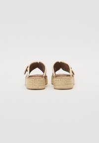 Deux sandales vues de dos avec des talons ouverts, des semelles en jute tissé, des tiges en tissu beige et une sangle avec une fermeture à bouton.