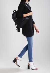 Frau, die in blauen Skinny-Jeans, einem schwarzen strukturierten Pullover und weißen, offenen High- Heels unterwegs ist und einen schwarzen Leder-Rucksack trägt.