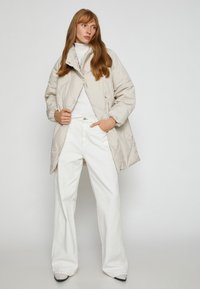 Lys beige polstret jakke med høj krave og trykknapper, parret med hvide brede jeans. Glat tekstur og afslappet design.