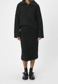 Sweatshirt noir, à texture duveteuse, avec un col zippé, associé à une jupe crayon assortie ; portée avec des chaussures à plateforme noires et des chaussettes.