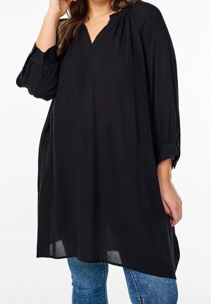 Kobieta ubrana w luźną czarną tunikę z długim rękawem i wycięciem przy dekolcie, w połączeniu z niebieskimi dżinsami, stoi na tle jednolitego białego tła.