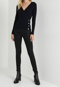 Tummansininen, v-pääntie ja ribbineulottu neulepaita, jossa on pitkät hihat, käärittävä malli ja kolme koristeellista valkoista nappia sivulla, yhdistetty mustiin housuihin.