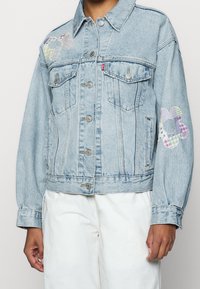 Giacca di jeans azzurro chiaro con toppe floreali ricamate in colori pastello sulle spalle, dotata di chiusura a bottoni e due tasche sul petto.