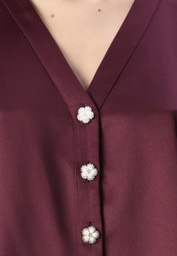 Burgundy blouse featuring a V-neckline and three floral-shaped buttons with pearl accents. Smooth texture and lightweight fabric.