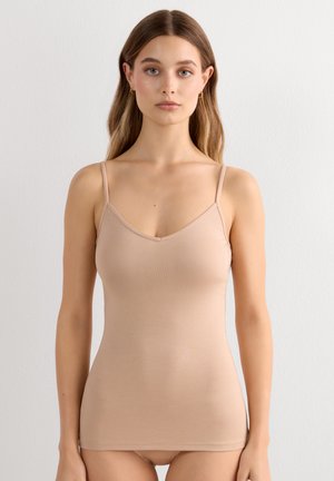 Ung kvinde med langt hår iført en beige ærmeløs top, står foran en ensfarvet hvid væg og vender fremad.