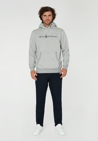 Sudadera gris con capucha y bolsillo frontal, con el logo "SAIL RACING". Combinada con pantalones azul marino y zapatillas blancas.