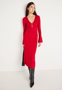 Rød ribbestrikket kjole med lange ermer, V-hals, dekorative frontknapper og vide mansjetter, sammen med svarte knelange spisse støvler.