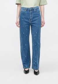 Modre kavbojke iz jeansa s krojem z ravnimi hlačnicami, z detajli v videzu obrabe in rahlim raztrganjem. Nošene z svetlo zeleno majico in črnimi čevlji.