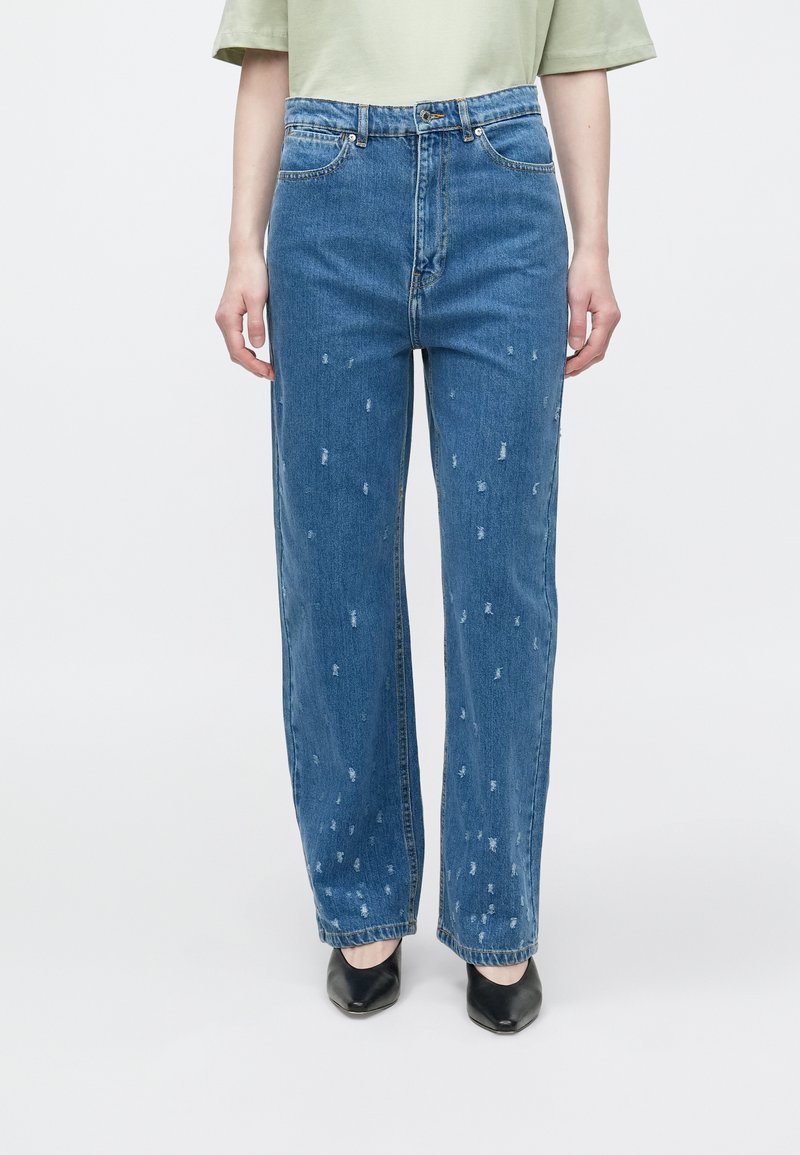 Modre kavbojke iz jeansa s krojem z ravnimi hlačnicami, z detajli v videzu obrabe in rahlim raztrganjem. Nošene z svetlo zeleno majico in črnimi čevlji.