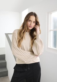 Pull en beige avec des manches bouffantes et côtelées associé à un pantalon noir ; texture douce, coupe décontractée et design minimaliste. Fond clair.