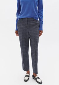 Pantalon gris à carreaux avec une coupe ajustée, souligné de lignes bleues et blanches. Porté avec un pull bleu et des mocassins noirs avec des détails blancs.