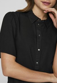 Black short-sleeve shirt with a structured collar, button details, and a smooth texture. Features subtle shoulder pleats.