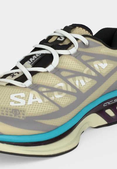 Αθλητικό παπούτσι σε μπεζ και μαύρο με λευκά κορδόνια, γκρι επικάλυψη, μπλε έμφαση στη μεσαία σόλα και λογότυπο "SACAI" στο πλάι και στη γλώσσα.