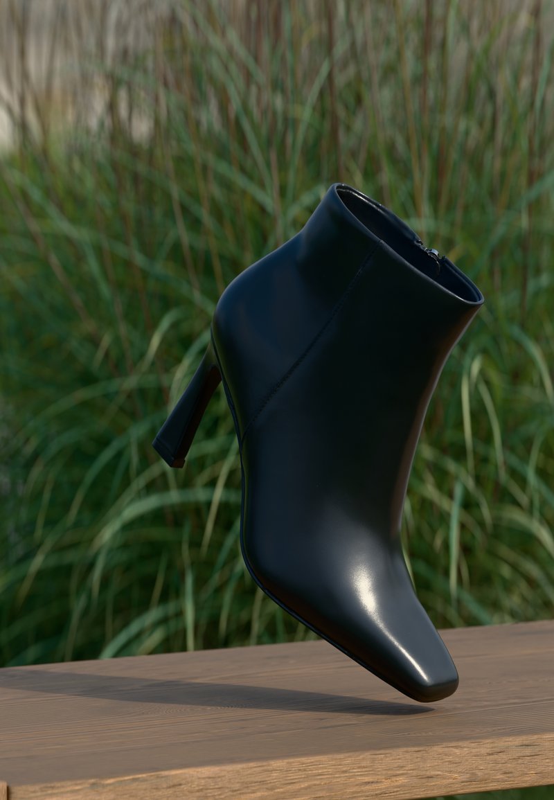Bota de cano curto preta em couro com bico fino e salto agulha; textura lisa e brilhante; possui um fecho lateral. O fundo sugere vegetação.
