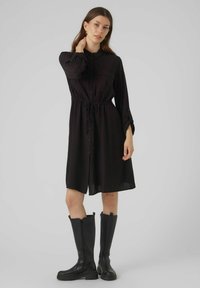 Rochie neagră până la glezne, cu talie ajustabilă, mâneci lungi, epauleți pe umeri și două buzunare pe piept. Asortată cu cizme negre până la genunchi.