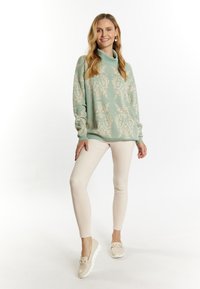 Light green sweater with an intricate cream pattern, cowl neck, and long sleeves; paired with fitted beige pants and light footwear.