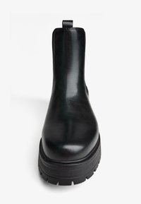 Czarny botek na kostkę wykonany z gładkiej syntetycznej skóry. Posiada zaokrąglony nosek, grubą gumową podeszwę z bieżnikiem oraz tylny uchwyt do zakładania.