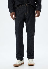 Vaqueros de mezclilla de color azul oscuro con corte recto, costuras naranjas y diseño de cinco bolsillos, acompañados de zapatillas blancas con acentos beige.
