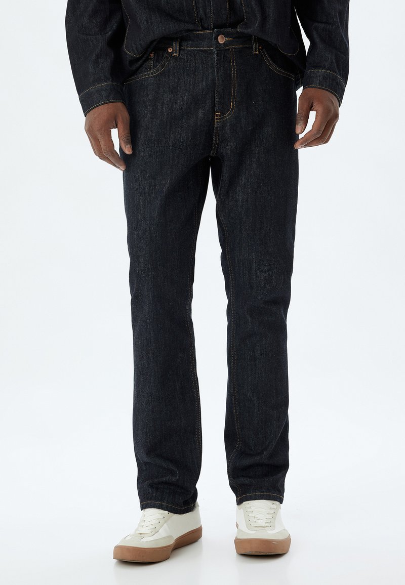 Vaqueros de mezclilla de color azul oscuro con corte recto, costuras naranjas y diseño de cinco bolsillos, acompañados de zapatillas blancas con acentos beige.