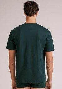 Ciemnozielona koszulka z krótkim rękawem, klasyczny krój, wykonana z gładkiej bawełnianej tkaniny. Charakteryzuje się luźnym fasonem i minimalnymi detalami w szwach.