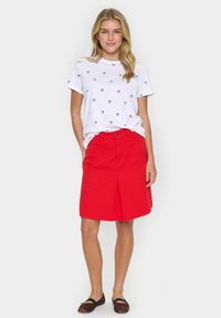 Balta T-kreklu ar melnu loku rakstu, apvienota ar sarkanu svārku līdz ceļiem, kuram ir priekšējais krokas. Modelis valkā brūnus zābakus.