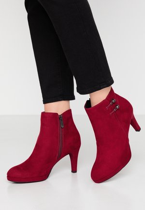 Bottes en daim rouge montant à la cheville avec fermeture éclair latérale et accents métalliques décoratifs, portées avec un pantalon noir ajusté.