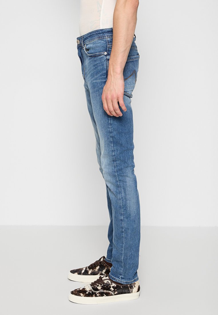 Męskie niebieskie dżinsy o slim fit, z przesunięciami kolorystycznymi i tradycyjnym pięciokomorowym fasonem, zestawione z butami o ciemnym wzorze.