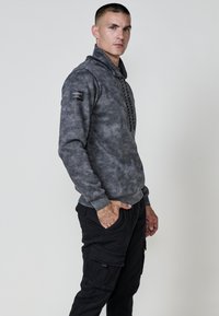 Hombre de pie de lado, vistiendo una sudadera gris con diseño de tie-dye y cuerdas negras, junto con pantalones cargo negros, con una mano en el bolsillo, sobre un fondo simple.