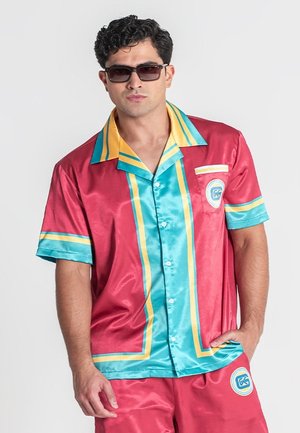 Muž so slnečnými okuliarmi v lesklej červeno-tyrkysovo-žltej košeli s gombíkmi a krátkych nohaviciach, stojaci s jednou rukou v vrecku.