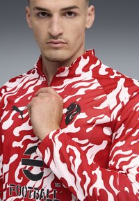 Rood-witte camouflagejas met een hoge kraag, voorzien van logo-accenten. Gemaakt van een gladde, lichte stof met een ritsluiting.