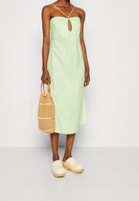 Hellgrünes Leinenkleid mit einem Schlüssel-Ausschnitt und dünnen Trägern, kombiniert mit einer tanfarbenen geflochtenen Tasche und cremfarbenen Clogs mit Holzsohlen.