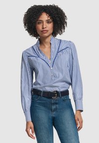 Blusa a maniche lunghe a righe blu e bianche con colletto a punta, dettagli con bottoni e cuciture blu a contrasto, abbinata a una cintura nera.