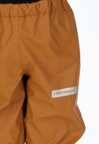 Waterproof oranje broek met een elastische tailleband. Heeft een zilveren "SWEDEMOUNT" logo op een witte rechthoekige patch. Glad, glanzend oppervlak.