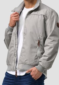 Man in een lichtgrijze sportjack met ritssluiting over een wit shirt, met een hand in de zak van zijn blauwe jeans, terwijl hij het jack op borsthoogte openhoudt.