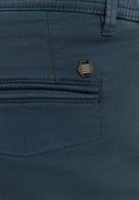 Donkerblauwe broek met een achterzak voorzien van een hexagonaal zwart en goud accent. Smooth structuur en subtiele stikdetails.