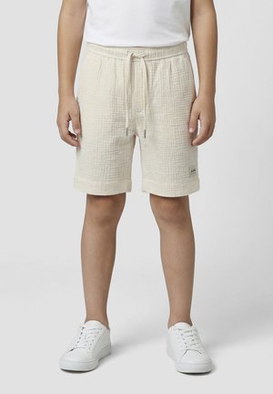 Enfant portant un short beige texturé avec cordon, un t-shirt blanc et des baskets blanches, debout les bras détendus sur les côtés.