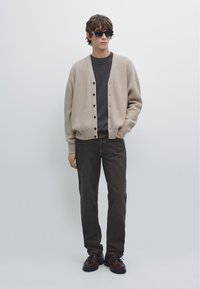 Jonge man met zwarte zonnebril, beige cardigan over een donkergrijs shirt, donkere jeans en bruine schoenen, staand tegen een lichte achtergrond.