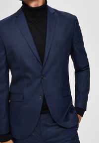 Mężczyzna w dopasowanym granatowym garniturze z zapiętą marynarką i czarnym golfem, z jedną ręką w kieszeni.