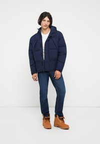 Chaqueta acolchada azul marino con capucha, con botones negros, llevada sobre una camisa blanca, acompañada de jeans azules y botas marrones con cordones.