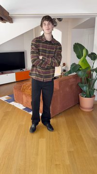 Kariertes Hemd in Dunkelgrün, Bordeauxrot und Gelb; schwarze Hose; schwarze Schuhe. Steht in einem Wohnzimmer mit einer Pflanze und einem braunen Sofa.