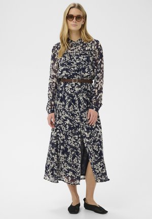 Kobieta w przezroczystej, granatowej sukience na długim rękawie w kwiaty, z brązowym paskiem, czarnymi balerinami i okularami przeciwsłonecznymi, stojąca na tle jasnej, jednolitej ściany.