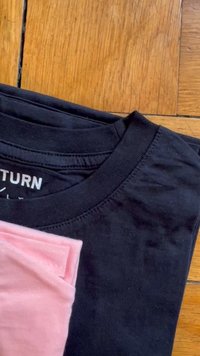 Čierne bavlnené tričko s okrúhlym výstrihom, vrstvené pod zloženou svetloružovou košeľou, odpočívajúce na drevenom povrchu.