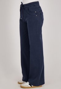 Marineblauwe wijde broek met trekkoord in de taille, voor- en achterzakken, gedragen met wit en zwarte sneakers op een neutrale achtergrond.