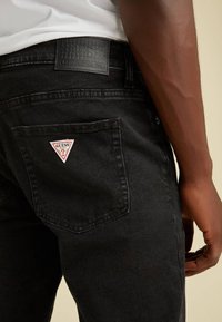 Jeans in denim nero con una patta in pelle nera testurizzata, logo a triangolo rosso sulla tasca posteriore e design a vestibilità slim.