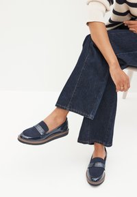 Navy slip-on kengät kiiltävästä lakasta, joissa on teksturoitu nauha ylhäällä ja kaareva ruskea pohja.